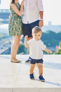 Adorable little boy making his first steps, parents are hugging in the background